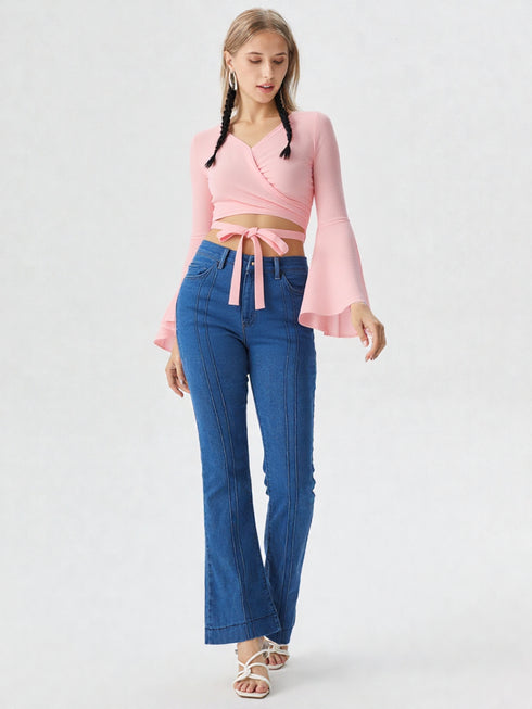 Woman wearing baby pink wrap front bell sleeve crop top with tie waist and blue jeans