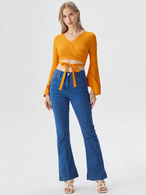 Woman wearing mustard yellow wrap front bell sleeve crop top with tie waist and blue flared jeans