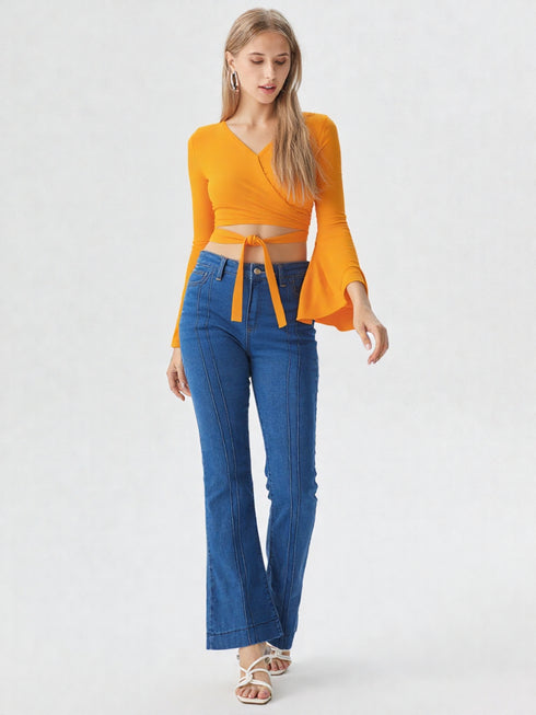 Woman wearing mustard yellow wrap front bell sleeve crop top with tie waist and blue flared jeans